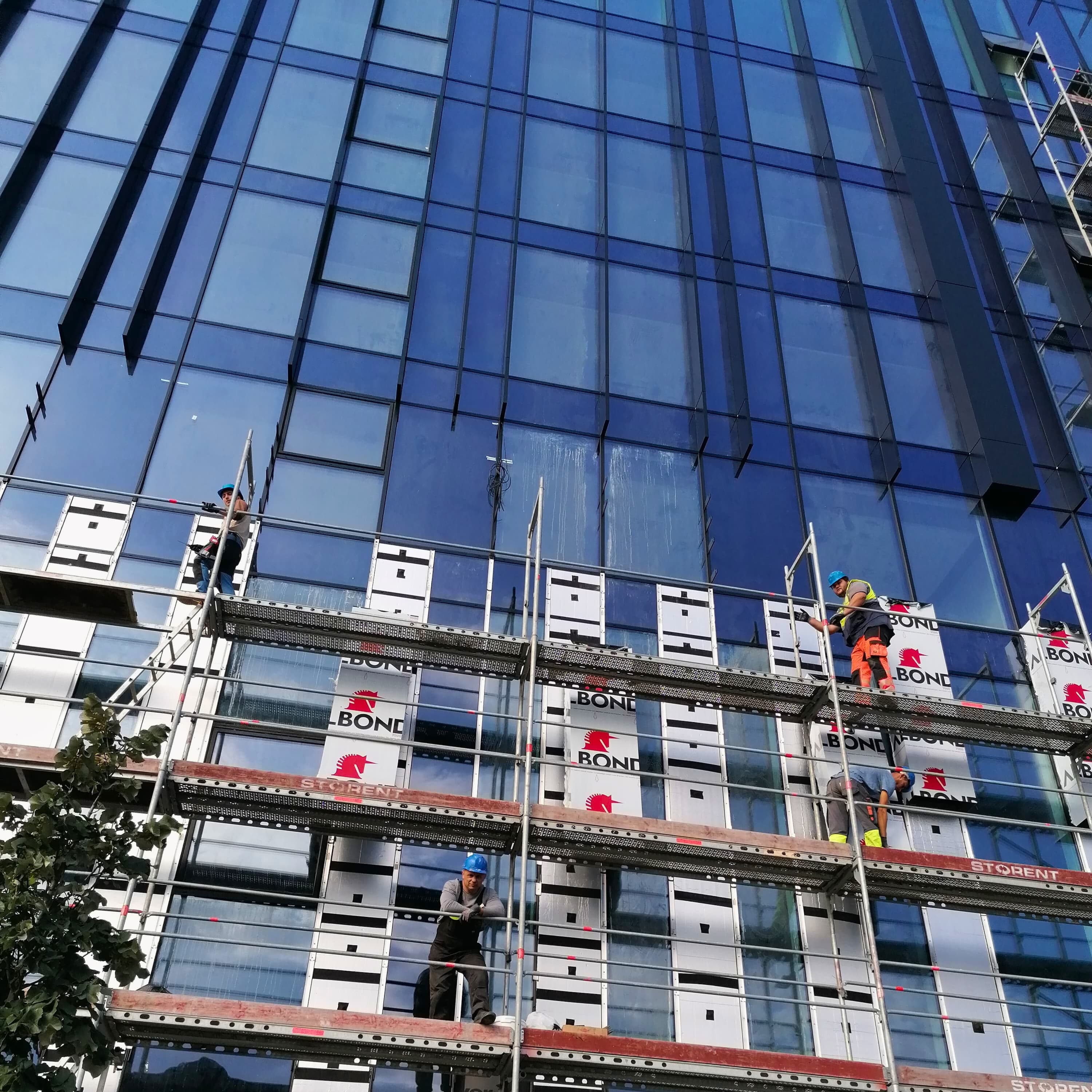 Darbininkų komanda su šalmais ir apsauginiais akiniais dirba ant pastolių prie aukšto pastato fasado.