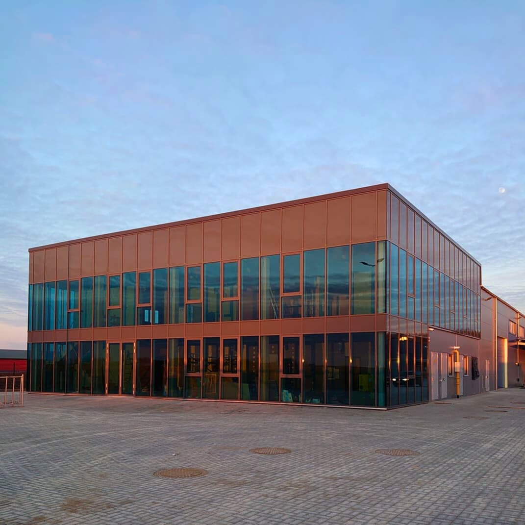 Gamyklos pastatas su plačiomis stiklo sienomis ir dideliu automobilių stovėjimo aikšteliu.