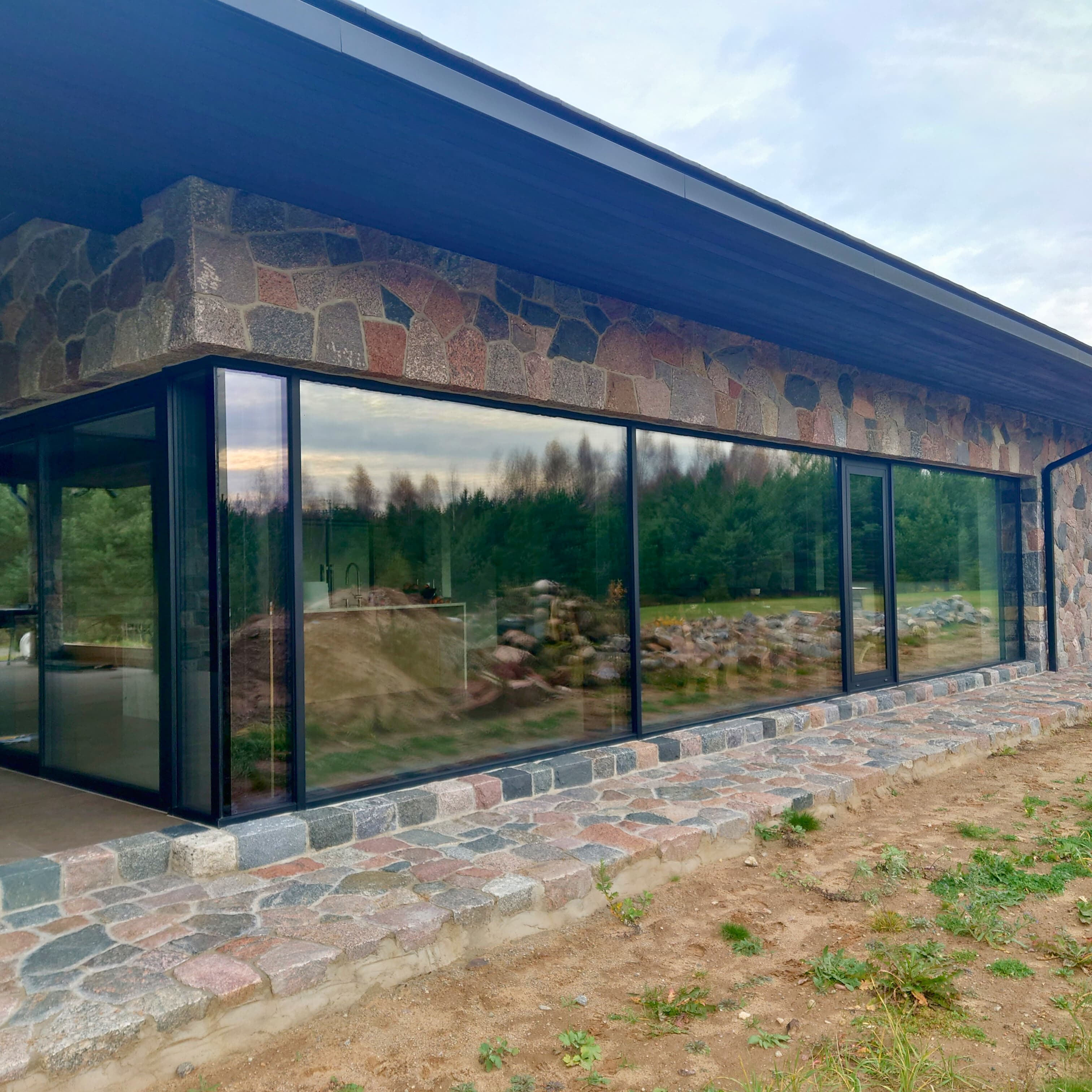 Šiuolaikinis namas su dideliais langais ir tekstūrinėmis akmens sienomis, derinantis modernią estetiką su gamtos elementais.